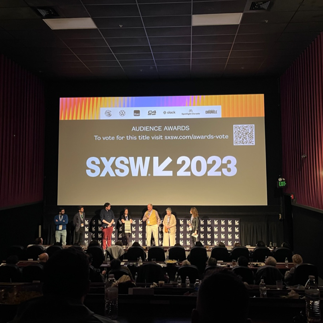 Seven adults stand on a stage with a large projector above them. Projector reads "Audience Awards. To vote for this title visit sxsw.com/awards-vote. SXSW 2023".